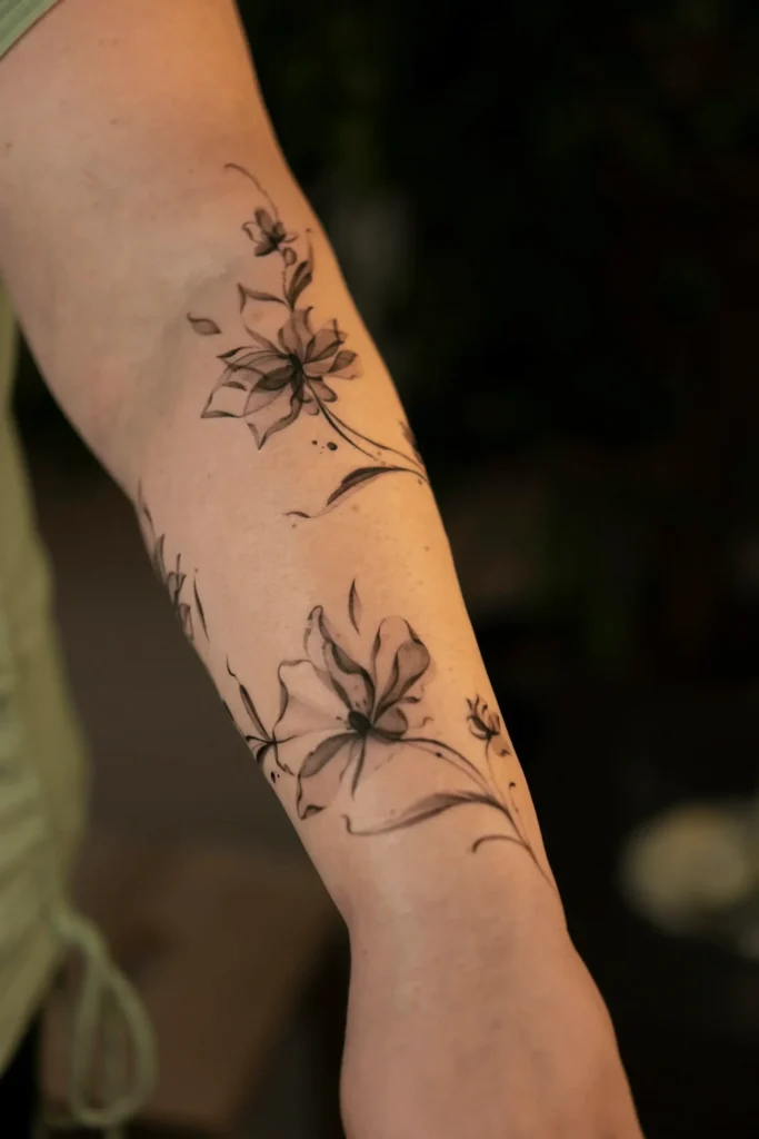 Delikatny, eteryczny tatuaż kwiatowy na przedramieniu z subtelnym cieniowaniem, wykonany w stylu fineline przez Wikę Olejnik w Częstochowie.