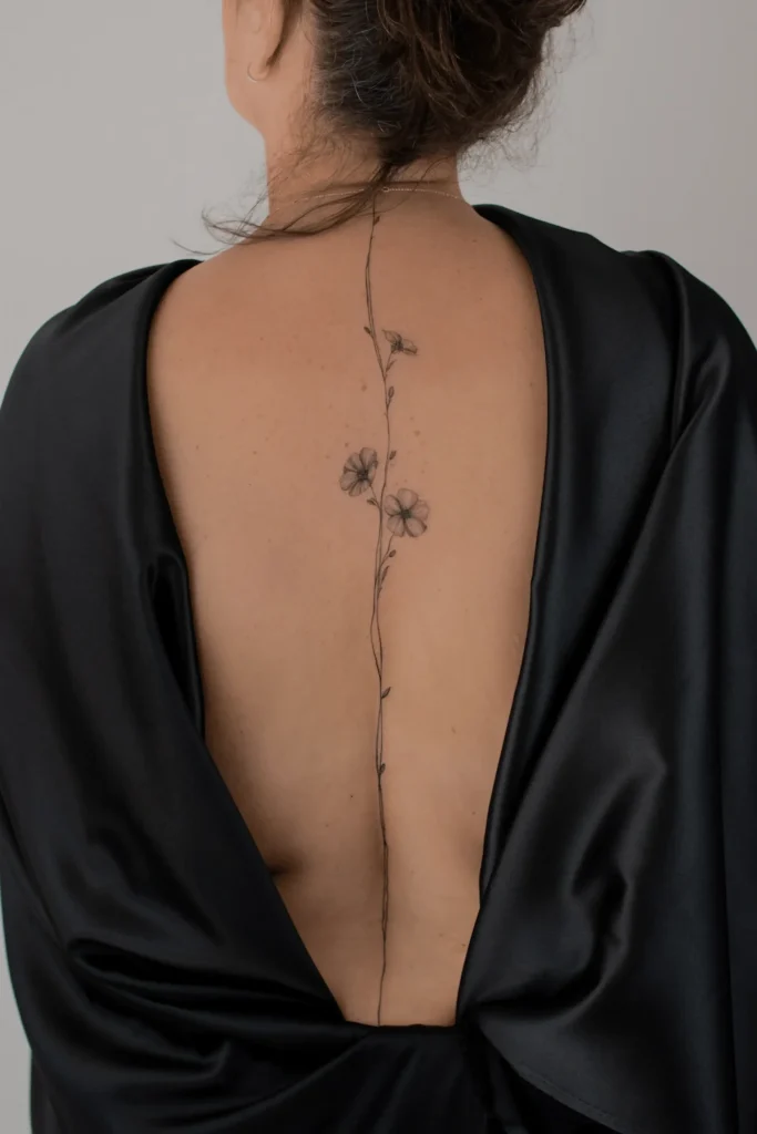 Bardzo delikatny, pionowy tatuaż botaniczny na kręgosłupie przedstawiający subtelne polne kwiaty, wykonany techniką cienkiej linii fineline przez Wikę Olejnik.