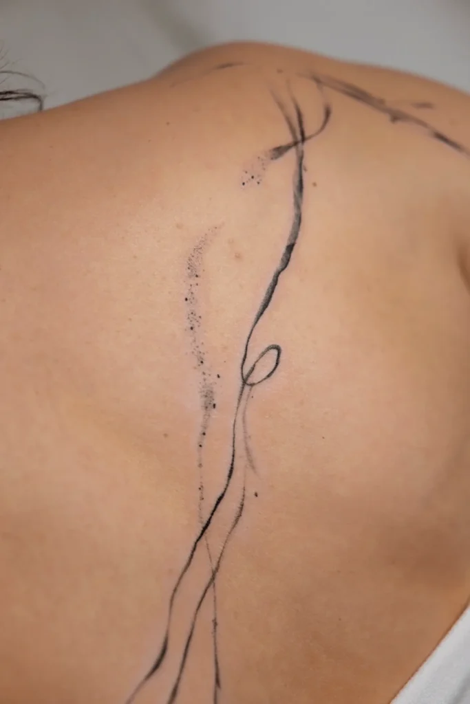 Minimalistyczny, abstrakcyjny tatuaż na plecach przedstawiający płynne, czarne linie i delikatny dotwork, podkreślający anatomię łopatki. Wykonanie: Wika Olejnik, Częstochowa.