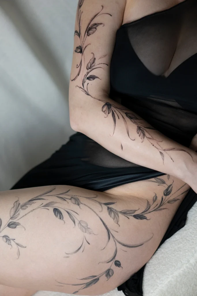 Duży, delikatny tatuaż botaniczny przechodzący przez ramię oraz biodro i udo, wykonany techniką cienkiej linii.