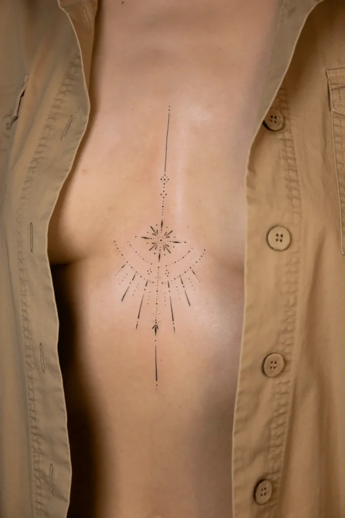 Minimalistyczny tatuaż fineline na mostku kobiety przedstawiający symetryczny ornament z centralną gwiazdą i kropkami dotwork. Wykonanie: Wika Olejnik, Częstochowa.