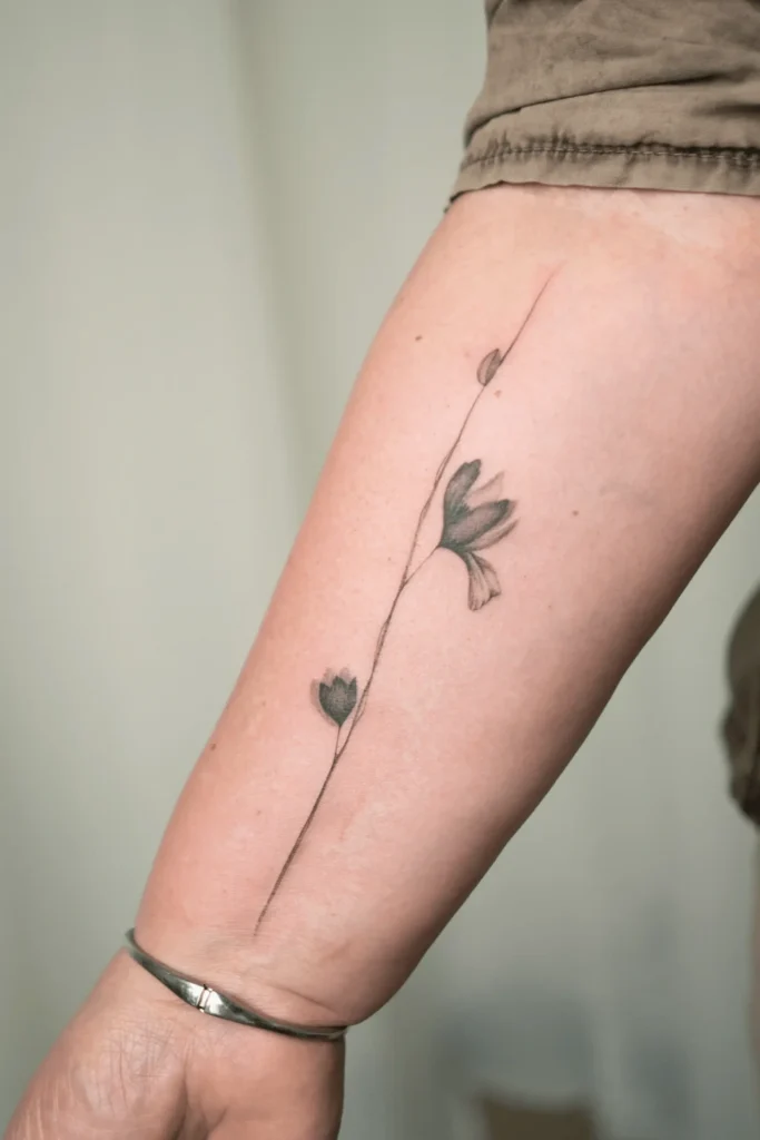 Minimalistyczny tatuaż fineline na przedramieniu przedstawiający pojedynczą, subtelną gałązkę z abstrakcyjnymi kwiatami. Wykonanie: Wika Olejnik, Częstochowa.