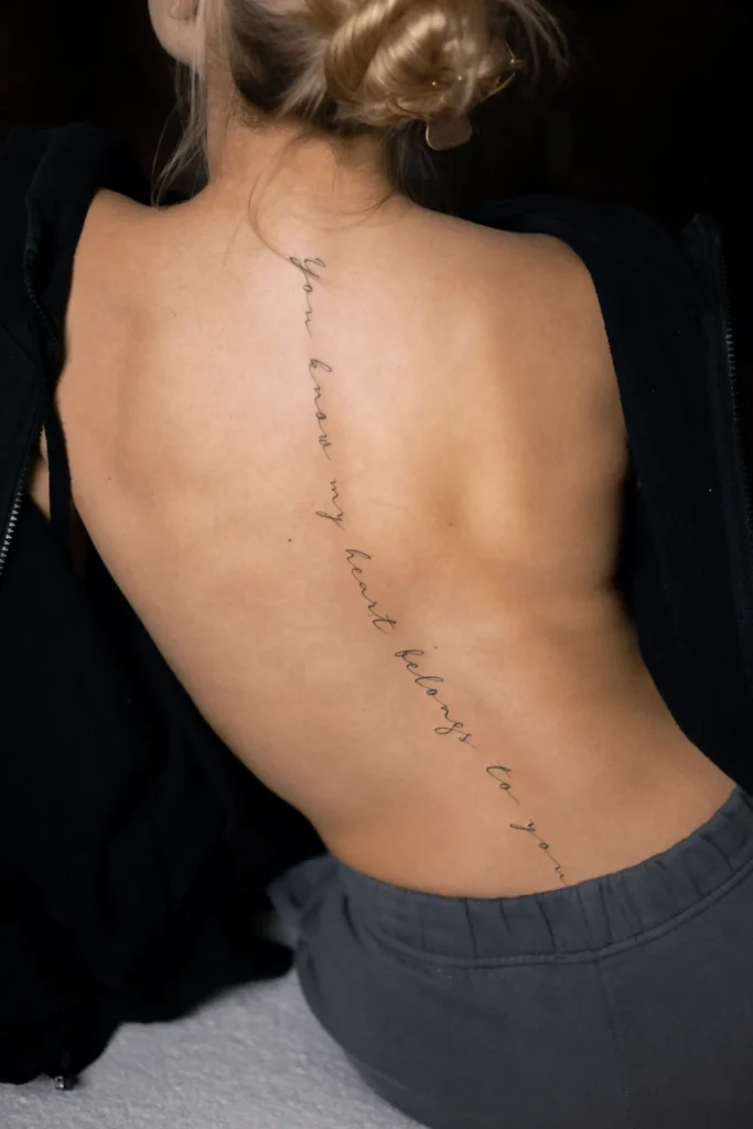 Pionowy tatuaż z napisem w stylu kaligrafii fineline wzdłuż kręgosłupa: 'You know my heart belongs to you'. Wykonanie: Wika Olejnik, Częstochowa.