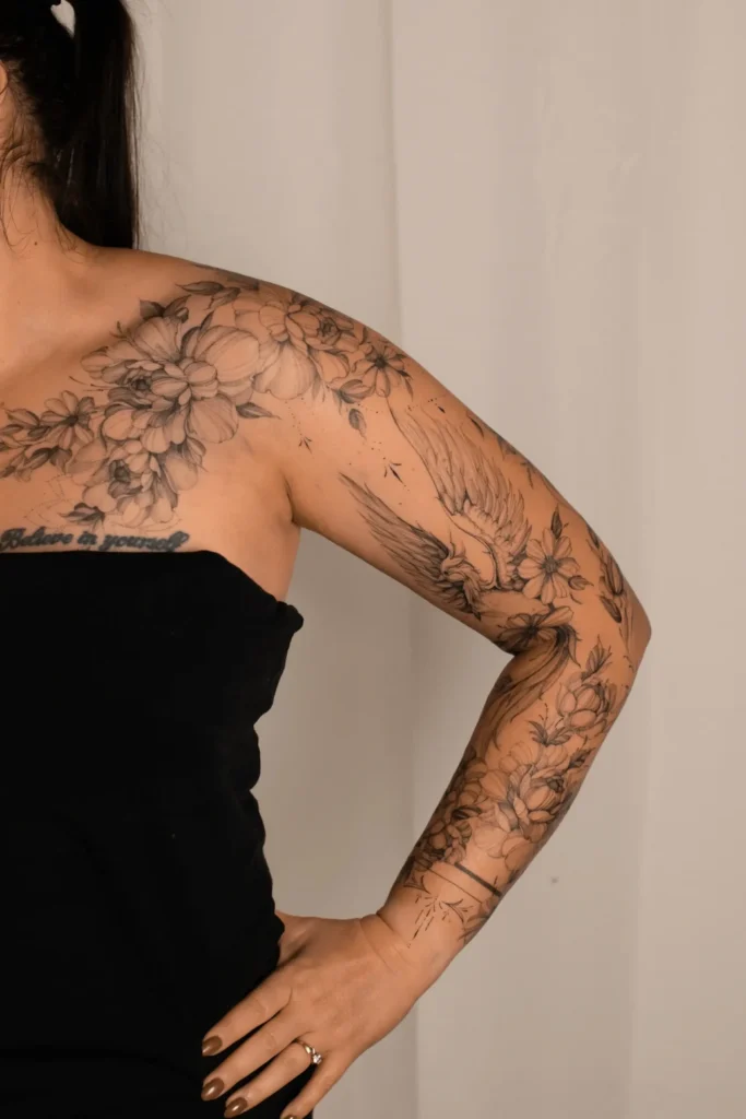Pełny tatuaż rękaw w stylu fineline na ramieniu kobiety, łączący bogatą kompozycję kwiatów (piwonie) z lecącym ptakiem. Wykonanie: Wika Olejnik, Częstochowa.