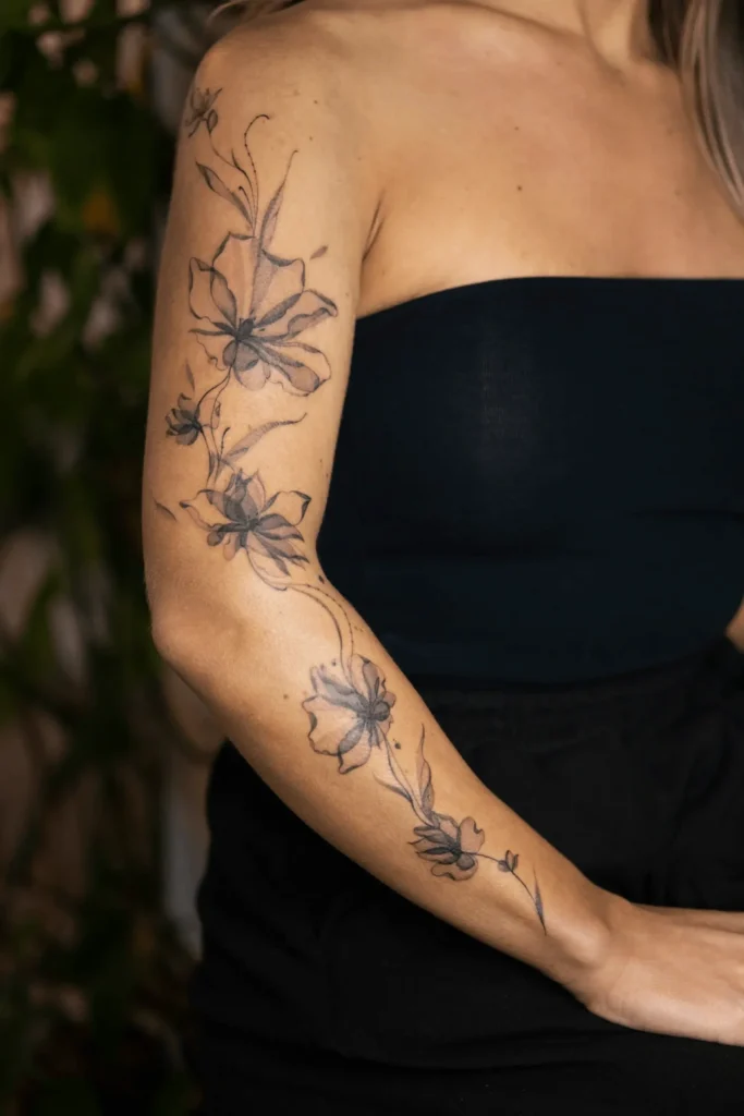 Kobiecy tatuaż typu rękaw przedstawiający eteryczne, prześwitujące kwiaty w stylu X-ray floral, wykonany techniką fineline przez Wikę Olejnik w Częstochowie.